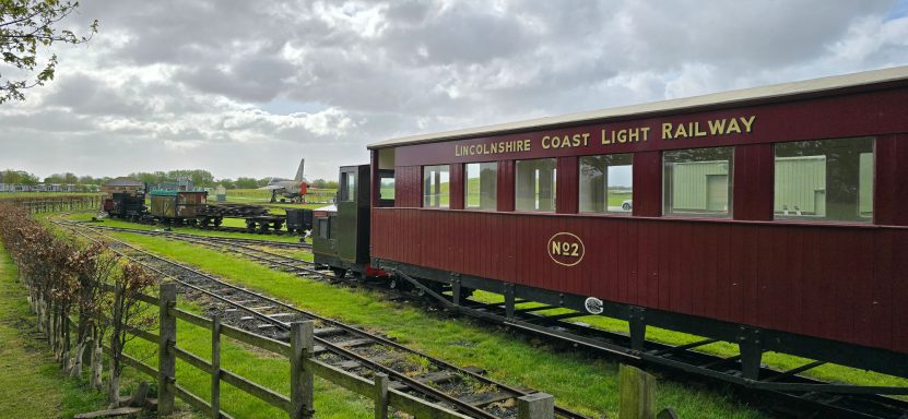 Red train carriage on a railway track with green fields and overcast sky in the background.