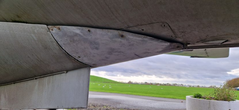 Underside view of a wing section showing a curved metal surface against a green field backdrop.